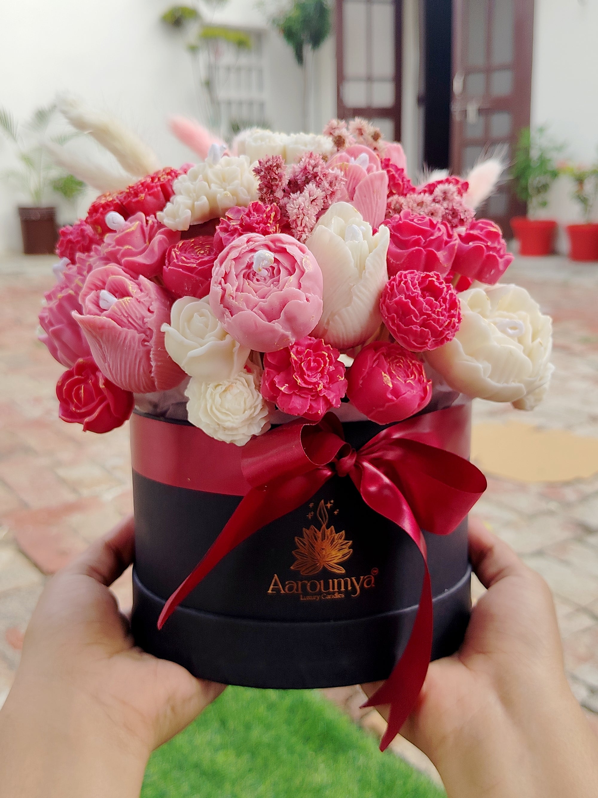 Floral candle Bouquet arrangement in a black box with a red ribbon held by hands, set against an outdoor background.